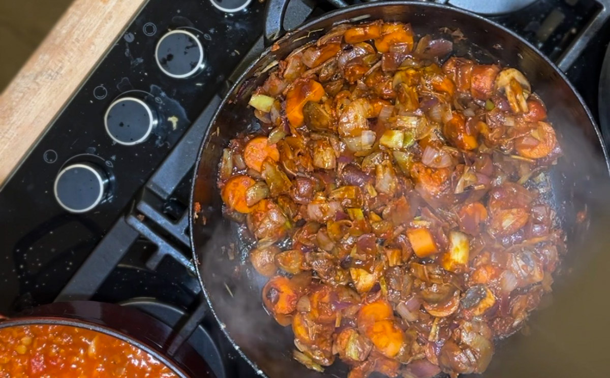 Vegane-Bratensuace-3 Gemüsepfanne mit Zwiebeln und Karotten auf einem Herd neben einem Topf mit Tomatensoße.