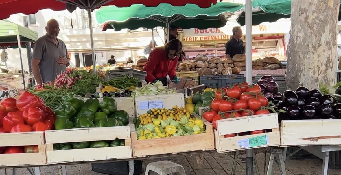 Vegane-Bratensauce-4 Marktstand mit frischem Gemüse: Paprika, Tomaten, Zucchini, Auberginen. Verkäufer und Kundin im Gespräch.