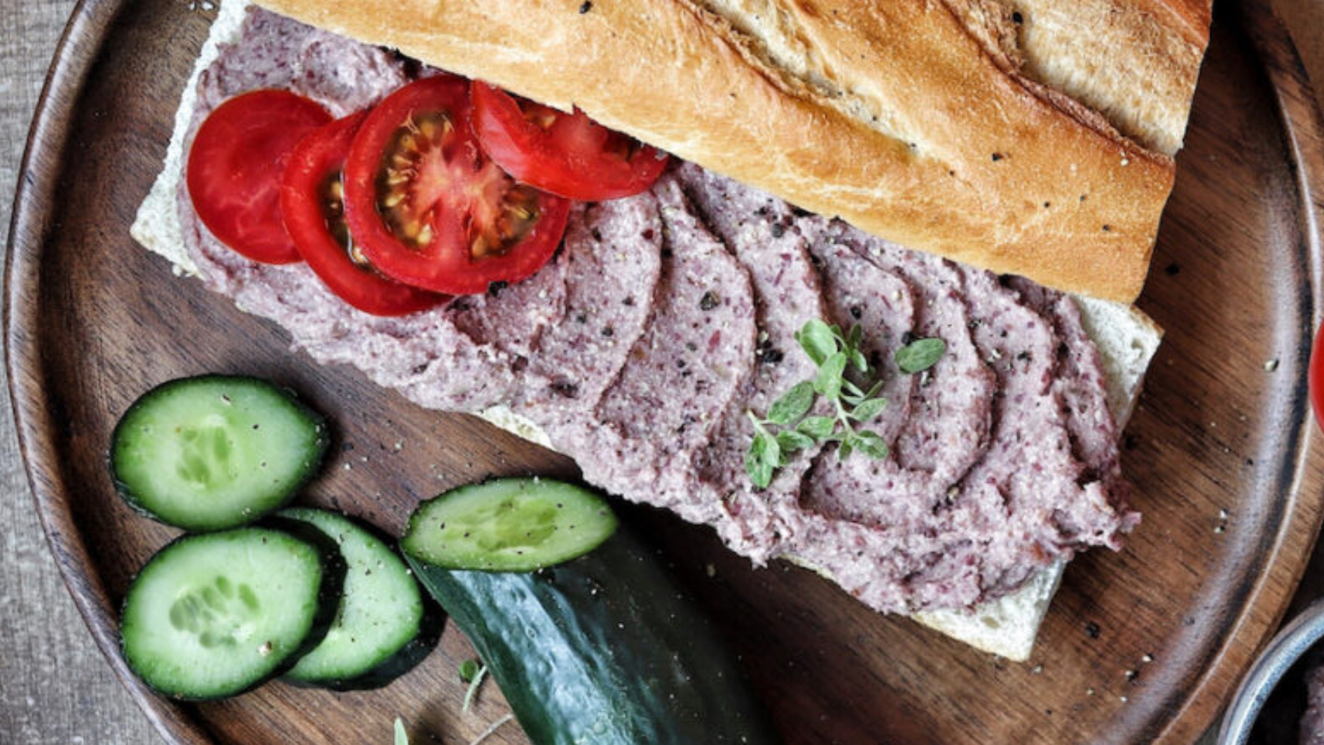 Baguette mit Aufstrich und Tomatenscheiben, daneben Gurkenscheiben auf Holzbrett.