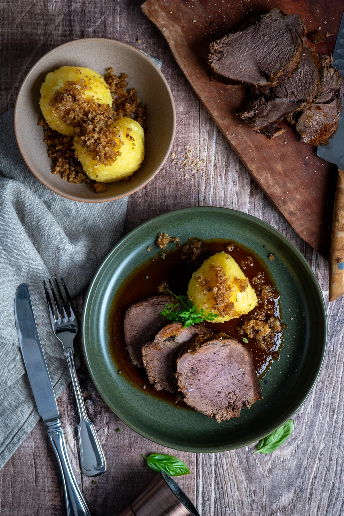 Teller mit Braten in Soße, Kloß und Semmelbröseln. Daneben: Messer, Gabel, Schüssel mit weiteren Klößen.