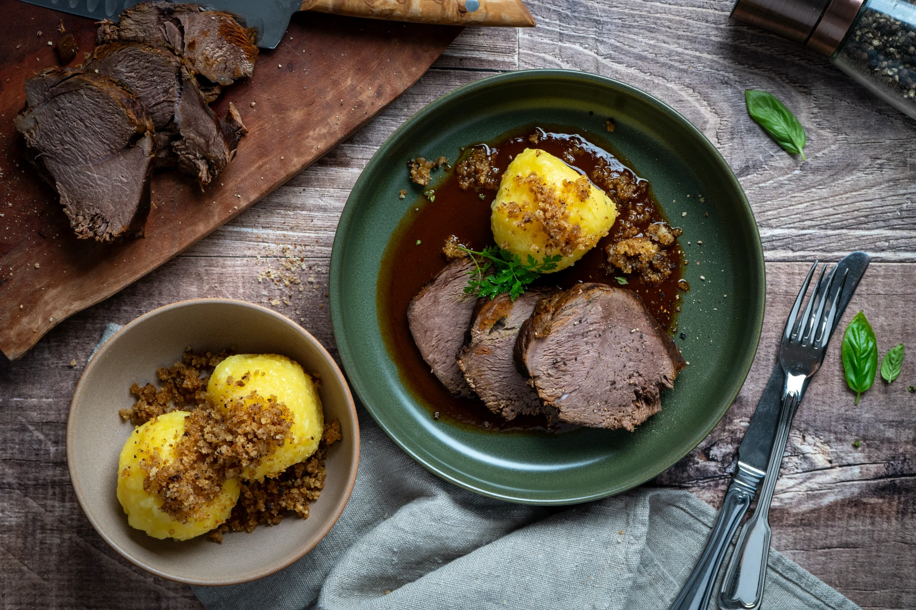 Gericht mit Scheiben Braten, Klößen und Soße auf grünem Teller, daneben Messer, Gabel, Brett mit Braten und Pfeffermühle.