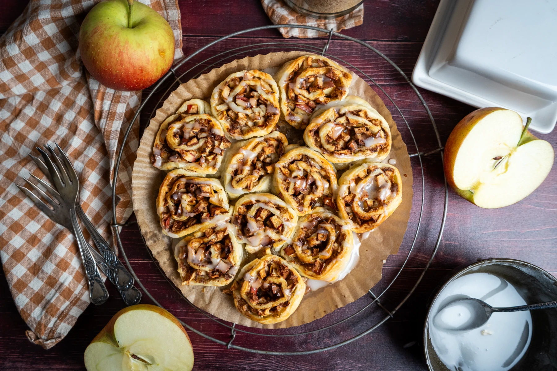 Apfelschnecken mit Zuckerguss auf Backpapier, umgeben von einem ganzen und einem halben Apfel, auf kariertem Tuch.