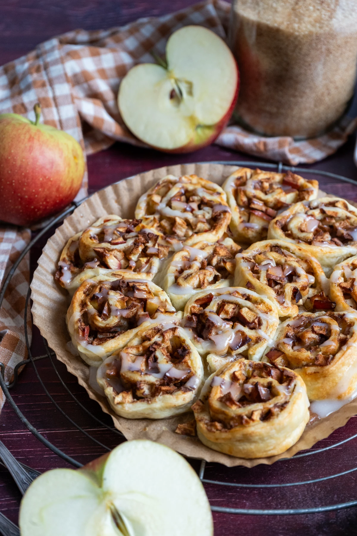 Frische Apfelschnecken mit Glasur auf einem Teller, umgeben von Äpfeln und braunem Zucker im Hintergrund.
