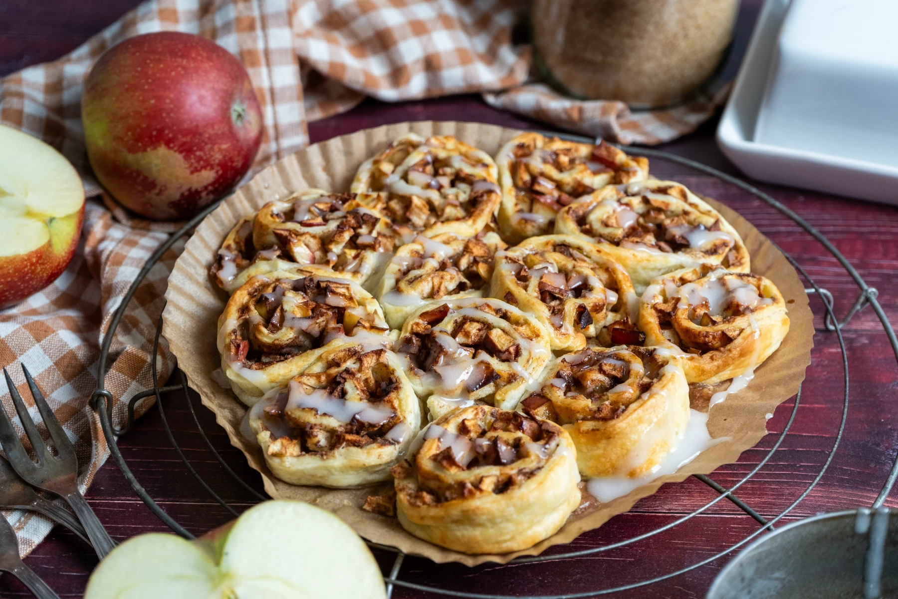 Zimtschnecken mit Apfelstücken und Zuckerguss auf einem Teller, umgeben von frischen Äpfeln und einem Geschirrtuch.