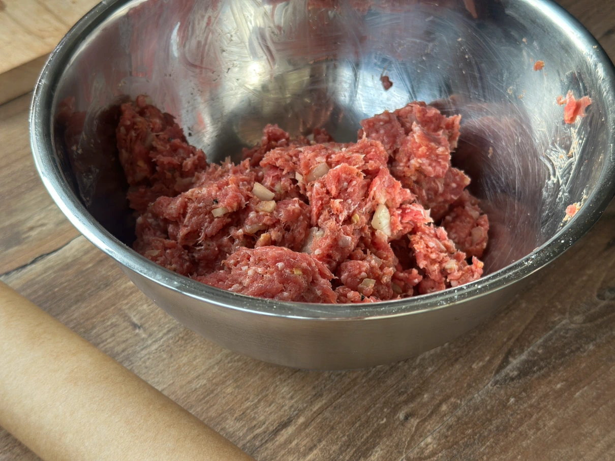 Edelstahl-Schüssel mit rohem Hackfleisch und Zwiebeln auf einem Holztisch.