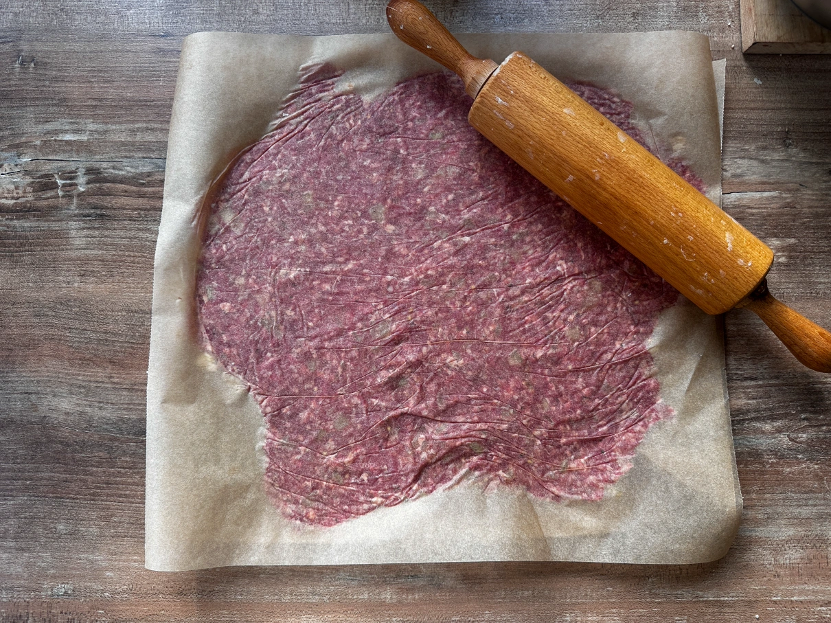 Ausgerollter Teig aus Hackfleisch auf Backpapier mit Holz-Nudelholz, auf Holztisch liegend.