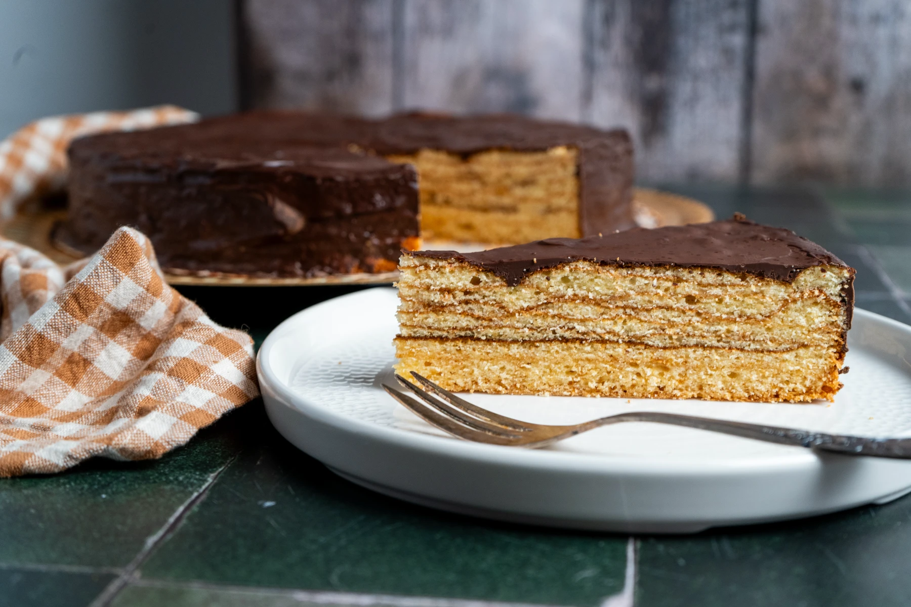 Ein Stück mehrschichtige Torte mit Schokoladenglasur auf Teller, daneben eine karierte Serviette. Hintergrund unscharf.