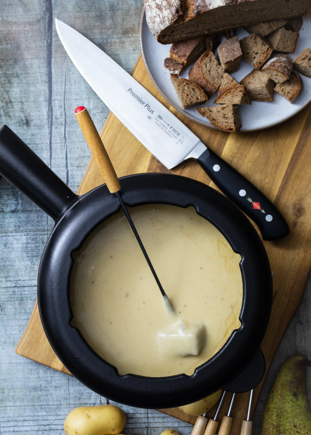 Fondue-Topf mit Käse, Gabel mit Brotstück, daneben Brotwürfel auf Teller und Messer auf Holzbrett.