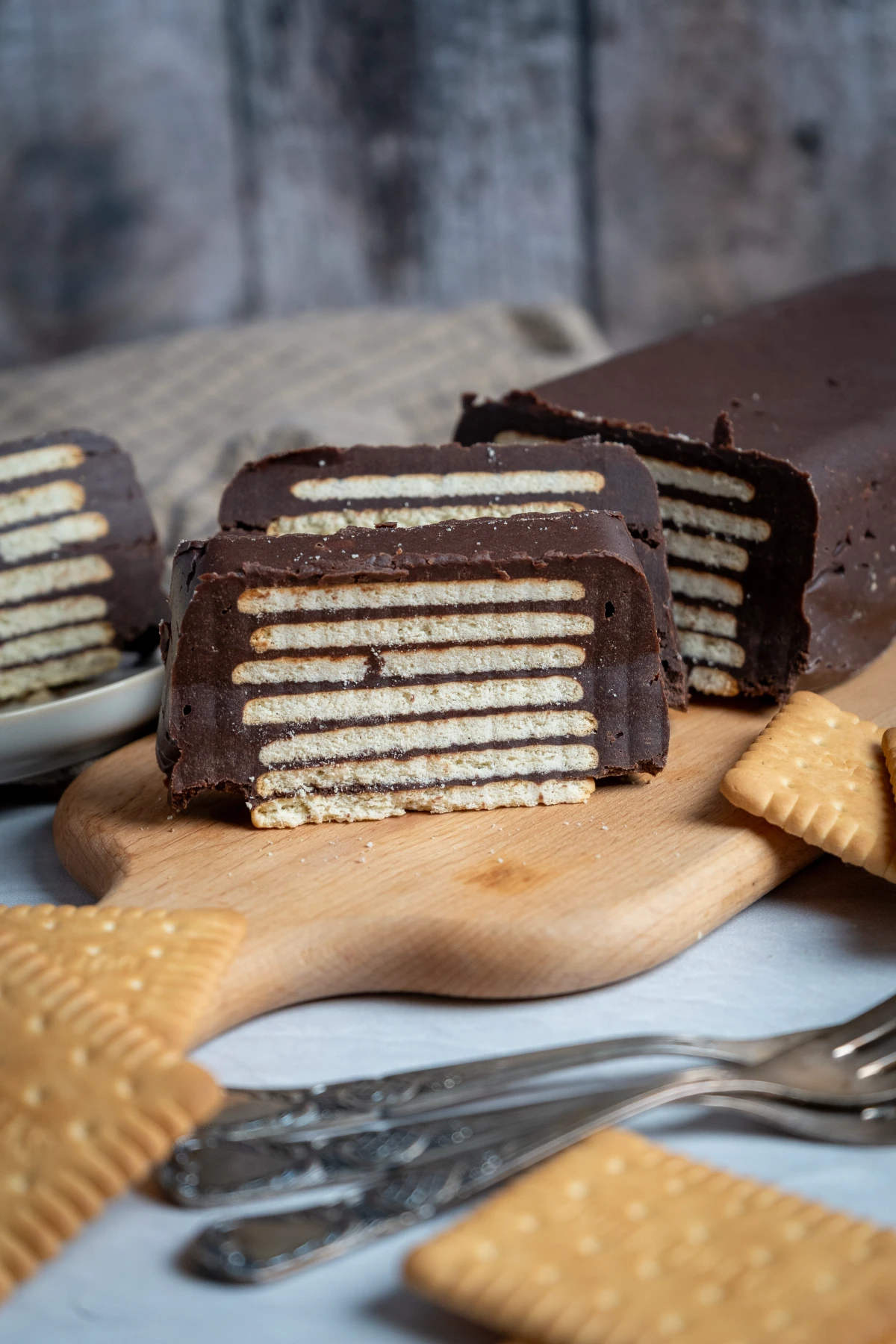 Schichttorte mit Keksen und Schokolade auf Holzbrett, zwei Stücke geschnitten, Keksstücke und Gabeln im Vordergrund.