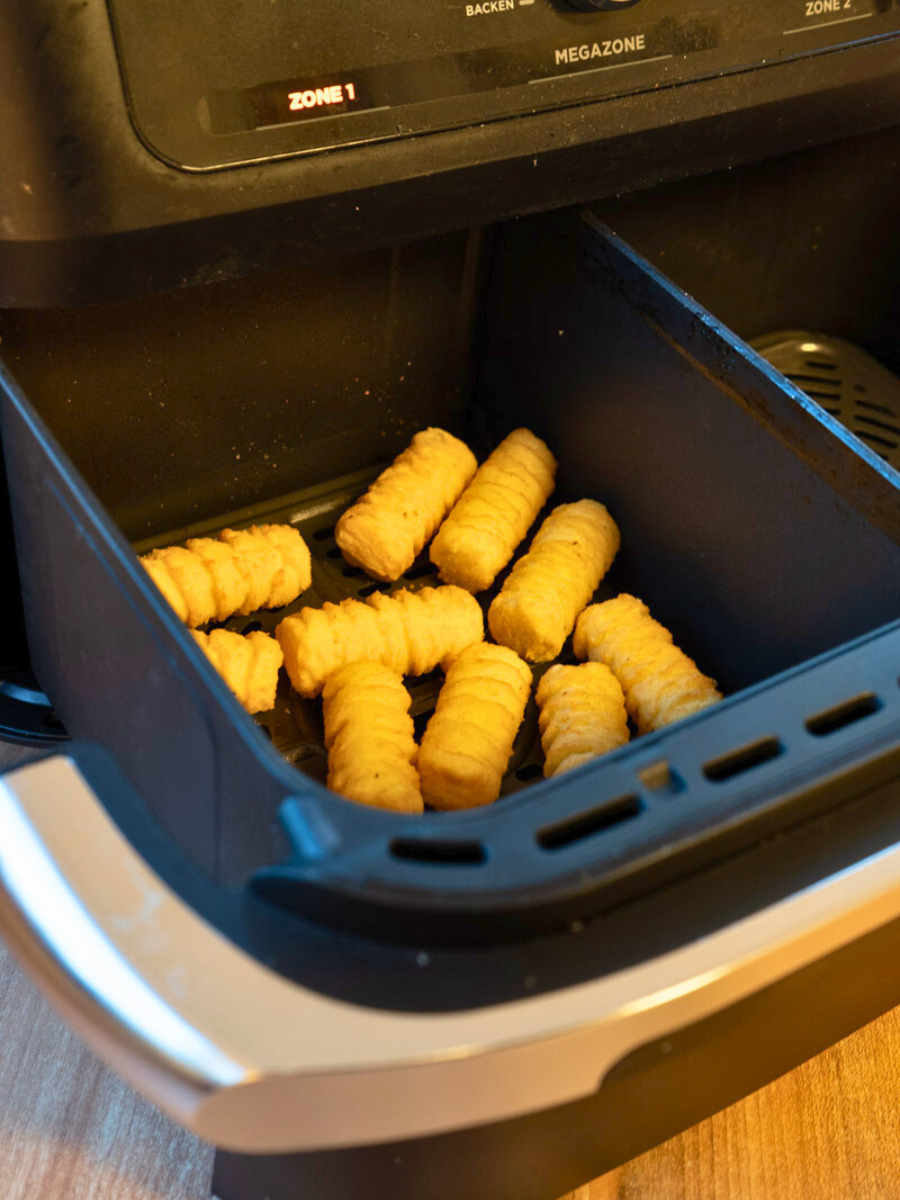Heißluftfritteuse-richtig-nutzen-04 Kroketten im Gittermuster liegen in der geöffneten Schublade einer Heißluftfritteuse auf einem Küchentisch.
