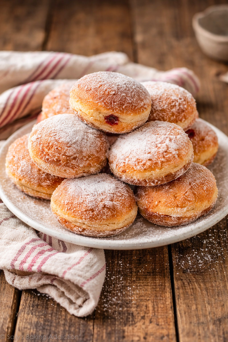 Ein Teller mit mit Zucker bestreuten Berlinern, gefüllt mit roter Marmelade, auf einem Holztisch.