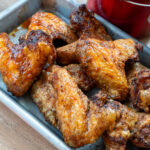 Gebratene Hühnerflügel auf einem Blech, knusprig gebräunt und saftig, neben einem roten Topf auf Holzuntergrund.