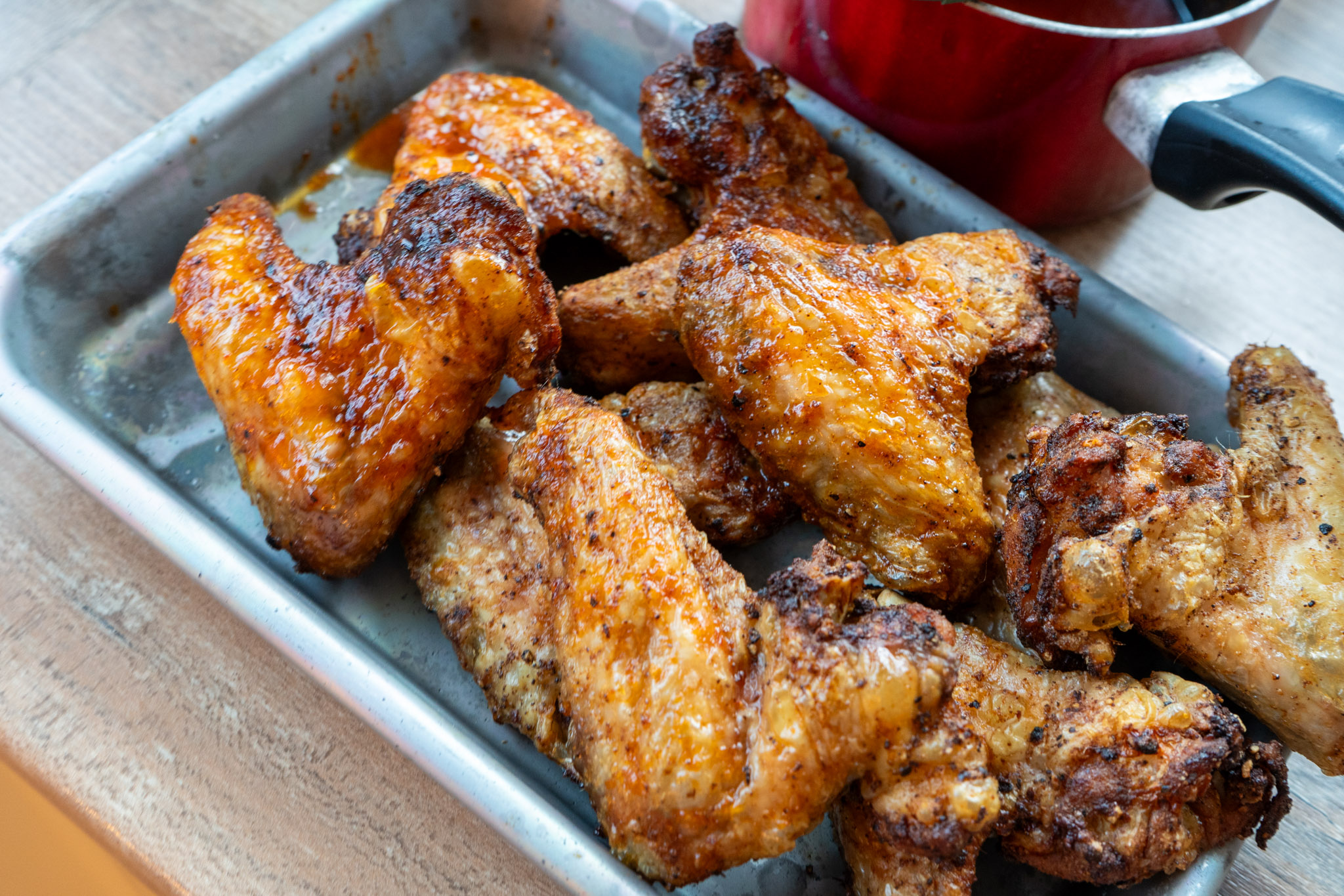 Buffalo-Chicken-Wings-01 Gebratene Hühnerflügel auf einem Blech, knusprig gebräunt und saftig, neben einem roten Topf auf Holzuntergrund.