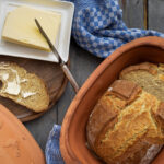 Frisch gebackenes Brot im Tontopf, daneben ein Holzbrett mit Butterbrot und Buttermesser auf blau-kariertem Tuch.