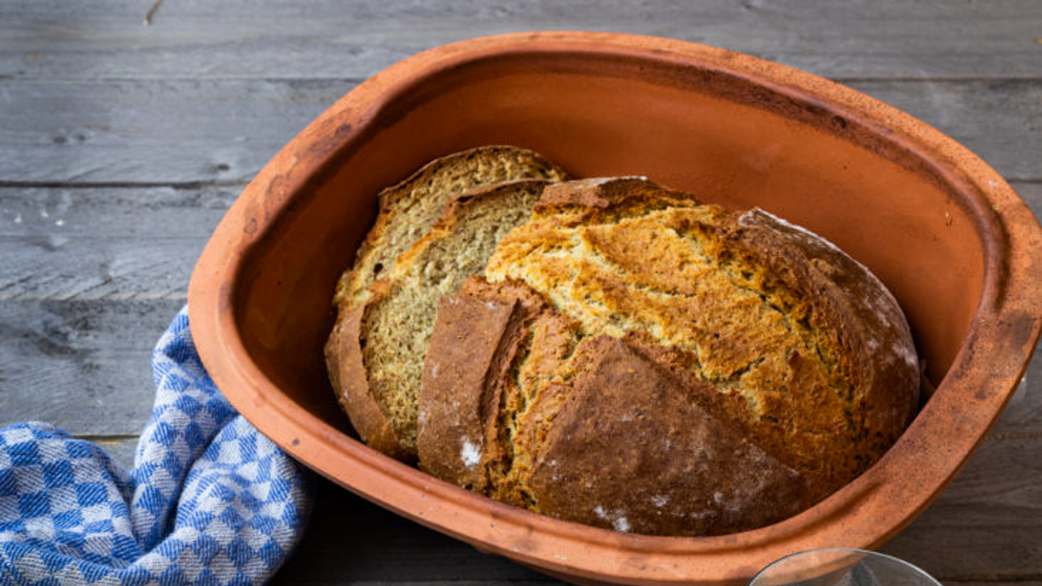 Brotlaib in einem runden, braunen Tontopf auf Holztisch mit blau-weißem Tuch.