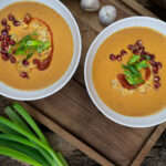 Zwei Schalen cremige Suppe mit Granatapfelkernen und Frühlingszwiebeln garniert, neben grünen Zwiebeln und Knoblauch.