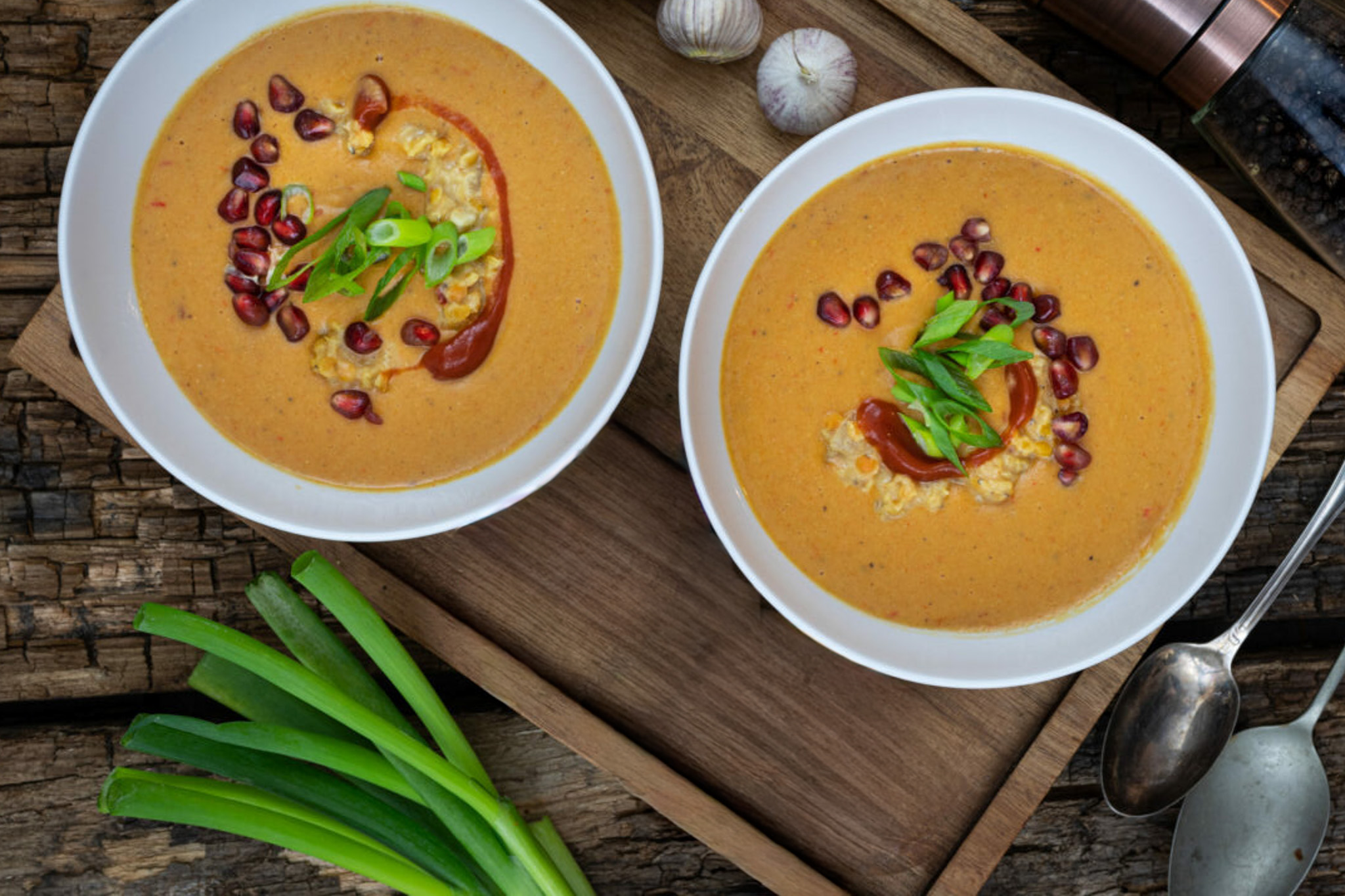 Curry-Linsen-Suppe-mit-Ingwer-01 Zwei Schalen cremige Suppe mit Granatapfelkernen und Frühlingszwiebeln garniert, neben grünen Zwiebeln und Knoblauch.