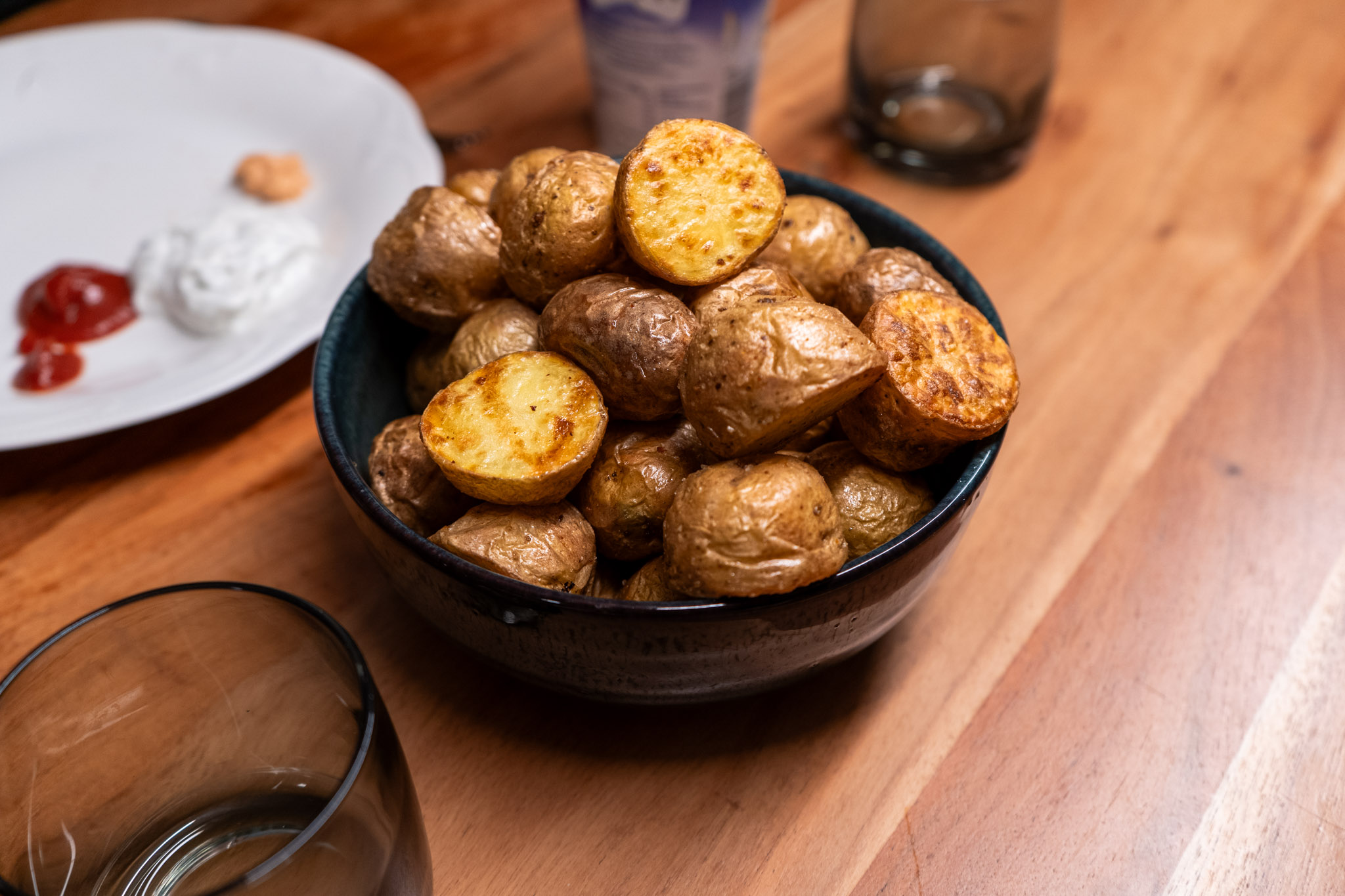 Schale mit gebackenen Kartoffeln auf Holztisch, daneben ein Teller mit verschiedenen Dips und ein leeres Glas.