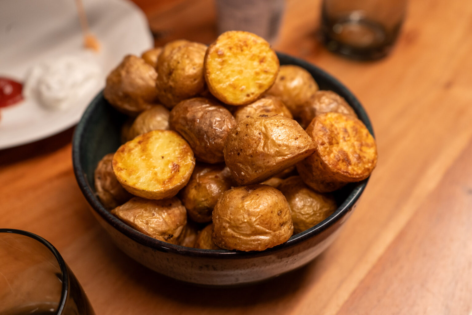 Schüssel mit knusprig gebackenen Kartoffelhälften auf Holztisch, mit Dip auf Teller im Hintergrund.