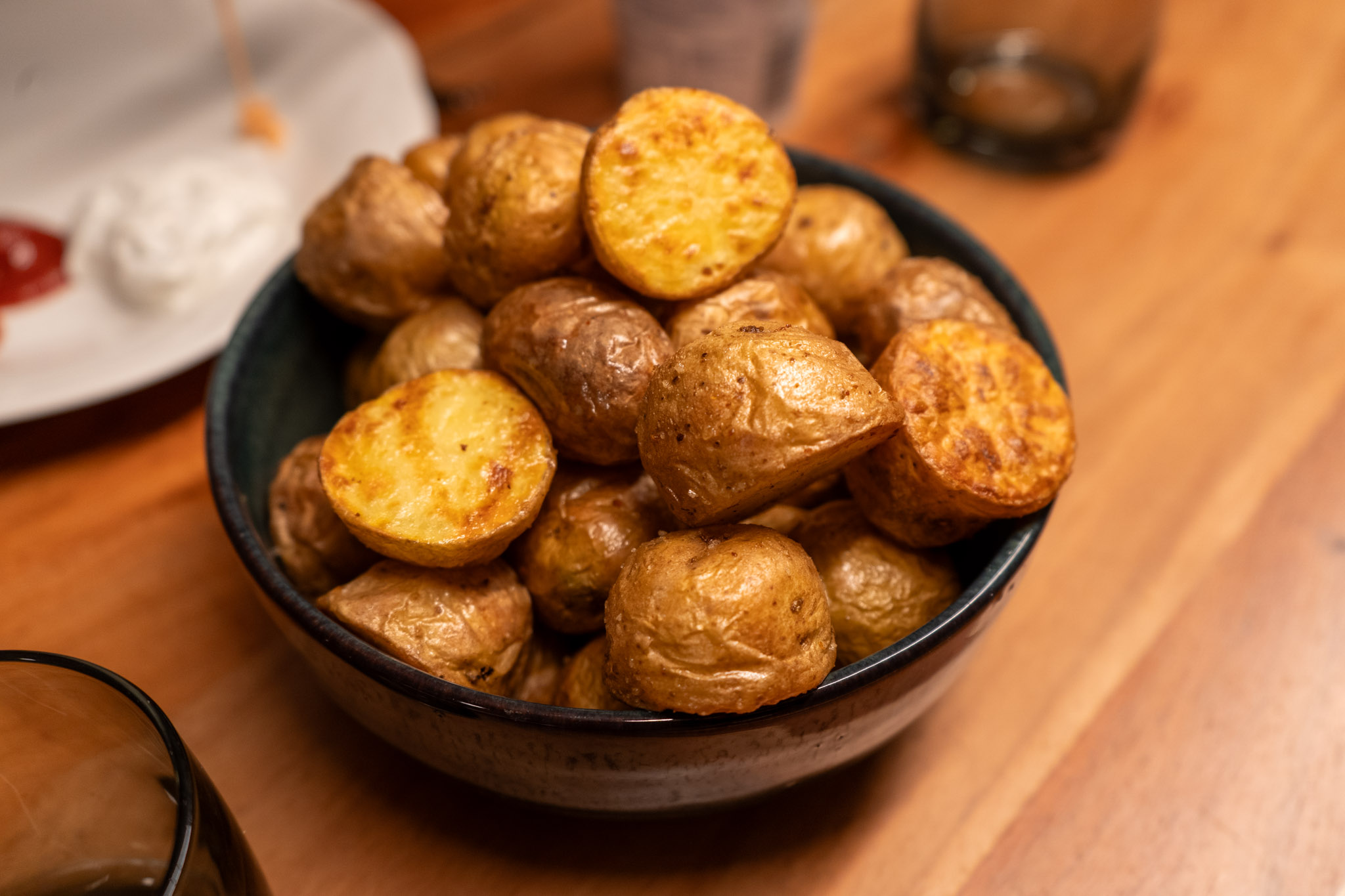 Drillinge-aus-der-Heißluftfritteuse-01 Schüssel mit knusprig gebackenen Kartoffelhälften auf Holztisch, mit Dip auf Teller im Hintergrund.