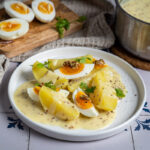 Gekochte Kartoffeln und Eier mit Senfsoße auf einem Teller, daneben ein Topf und ein Schneidebrett mit Eierhälften.