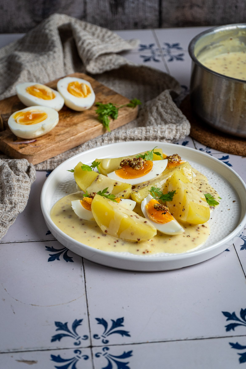 Eier-in-Senfsauce-02 Gekochte Eier und Kartoffeln in Senfsoße auf einem Teller, mit Petersilie garniert. Hintergrund: Topf, Holzbrett.
