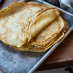 Ein Stapel frisch gebackener Crêpes auf einem Metallblech auf einem Holztisch.