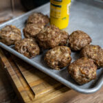 Fleischbällchen auf einem Backblech neben einer gelben Senf-Flasche, auf einem hölzernen Schneidebrett.