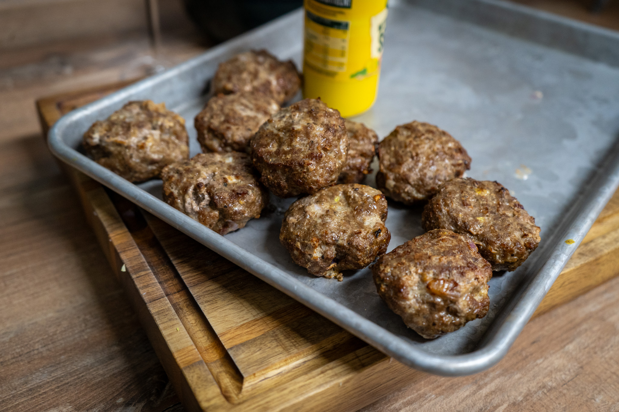 Frikadellen-aus-der-Heißluftfritteuse-01 Fleischbällchen auf einem Backblech neben einer gelben Senf-Flasche, auf einem hölzernen Schneidebrett.