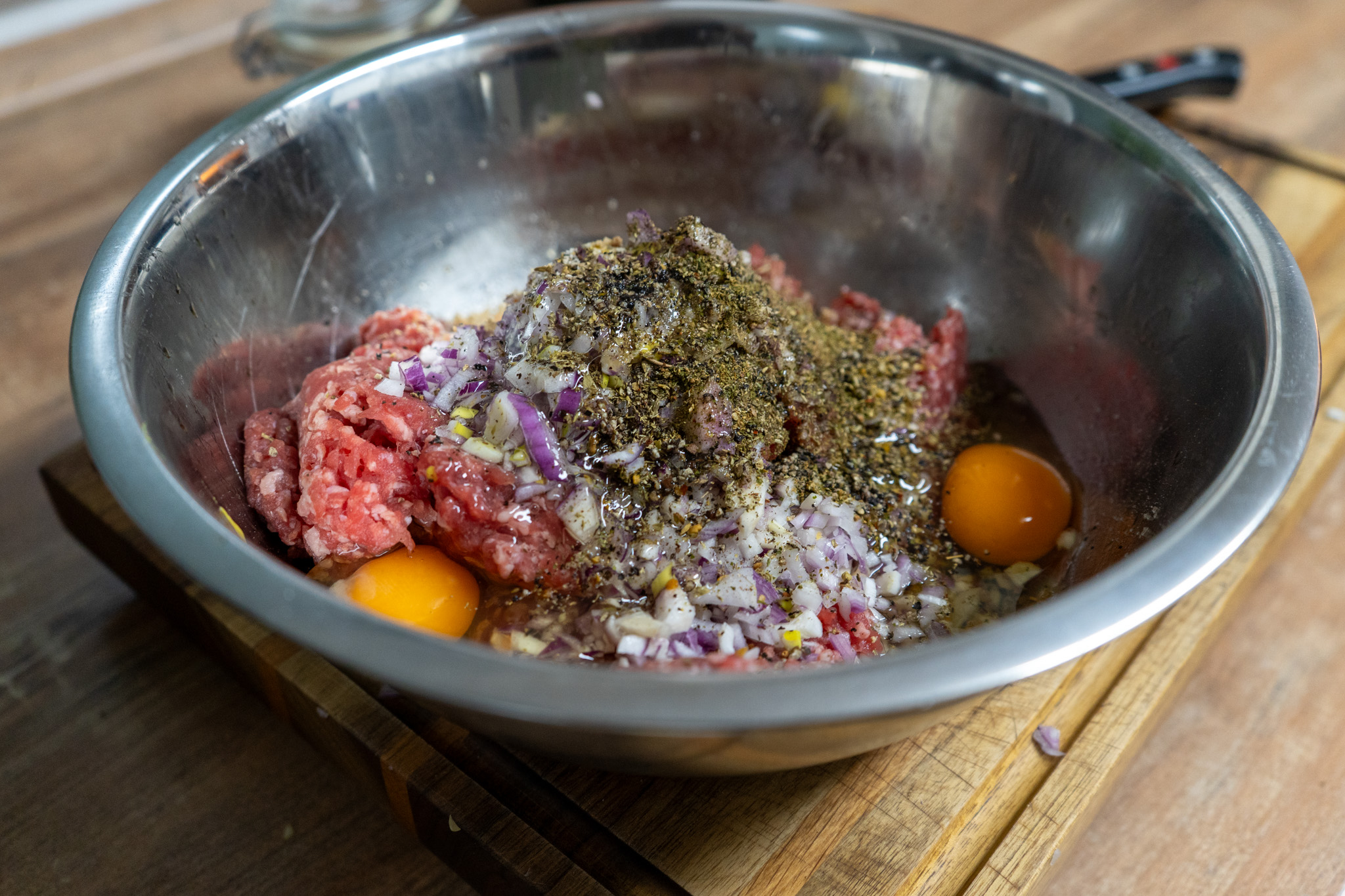 Frikadellen-aus-der-Heißluftfritteuse-06 Edelstahlschüssel mit rohem Hackfleisch, Eiern, gehackten Zwiebeln und Gewürzen auf einem Holzbrett.