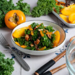 Salatschale mit Grünkohl, Pfirsichspalten und Nüssen auf Tisch. Umgeben von frischen Zutaten und Küchenutensilien.