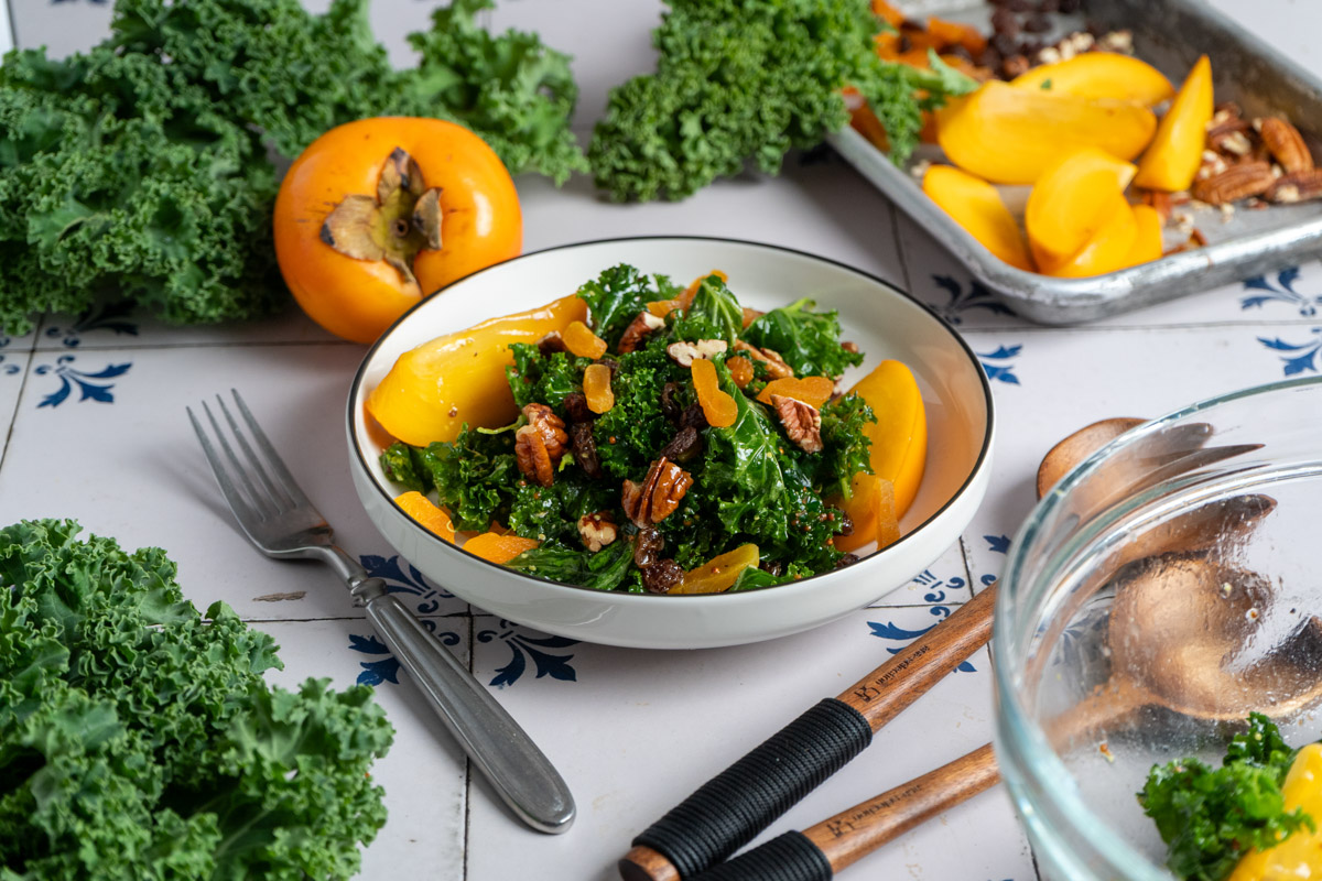 Salatschüssel mit Grünkohl, Persimonen, Nüssen und getrockneten Aprikosen. Frische Zutaten und Utensilien umgeben die Schale.