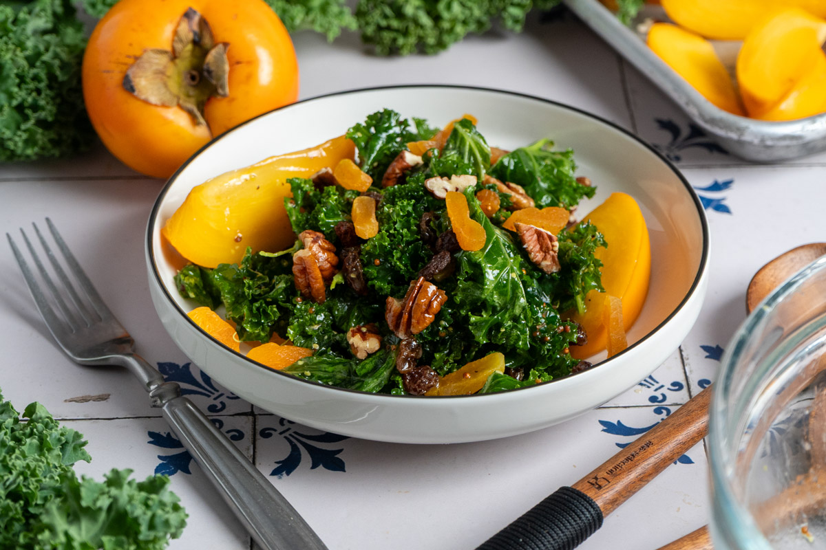Schale mit Grünkohl-Salat, Pekannüssen und getrockneten Früchten; daneben eine frische, orange Kakifrucht.