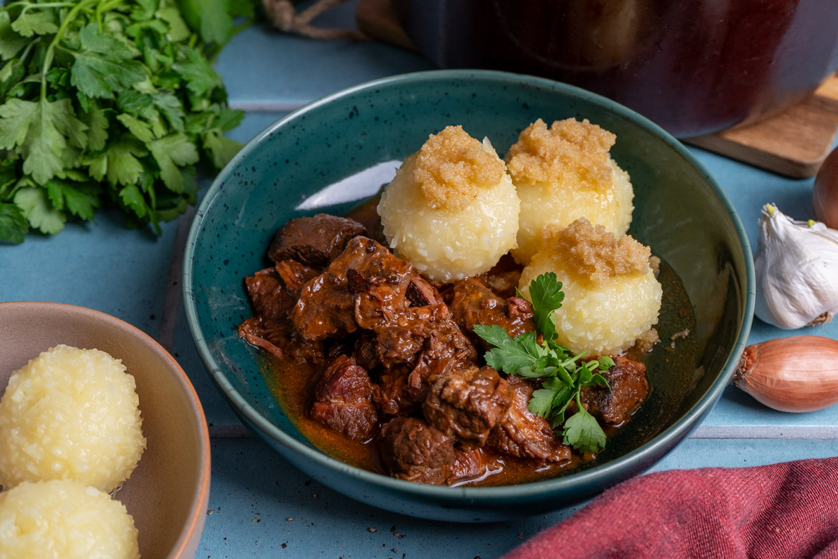 Grüner Teller mit Gulasch, drei Kartoffelknödeln, Petersilie und Bröseln. Daneben Knoblauch, Petersilie und Schalotten.