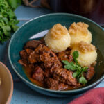 Schüssel mit Rindergulasch, drei Klößen mit Bröseln, garniert mit frischer Petersilie. Petersilie, Knoblauch und Zwiebel daneben.