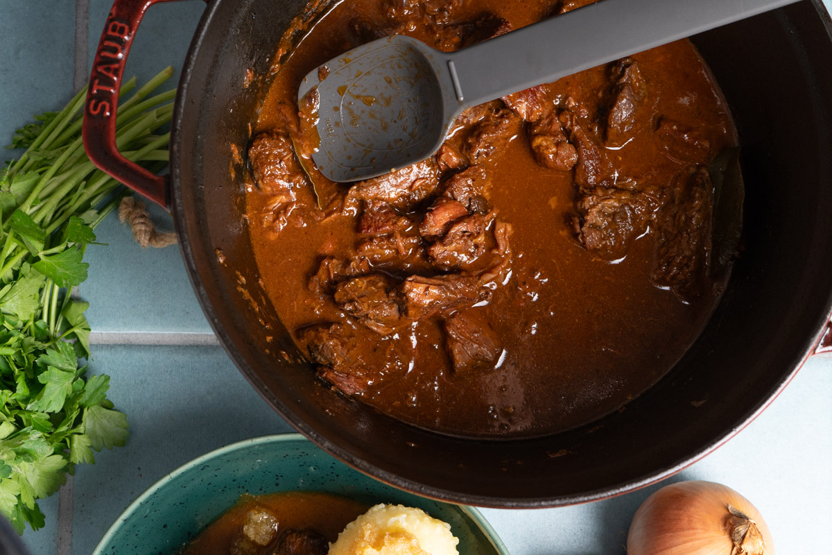Rindergulasch in einem Topf mit Löffel, daneben frische Petersilie, ein Teller mit Kartoffelpüree und eine Zwiebel.