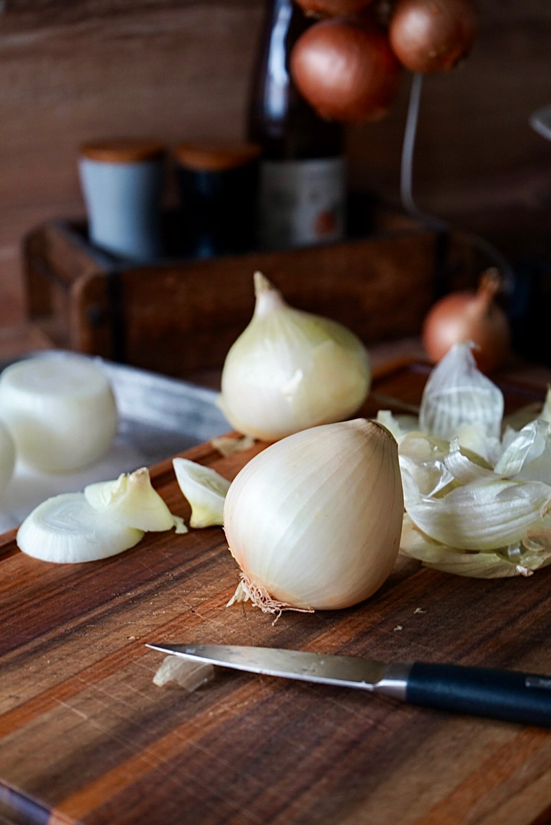 Zwiebel auf Schneidebrett mit Schälresten und kleinem Messer, Hintergrund mit weiteren Zwiebeln und Gewürzen.