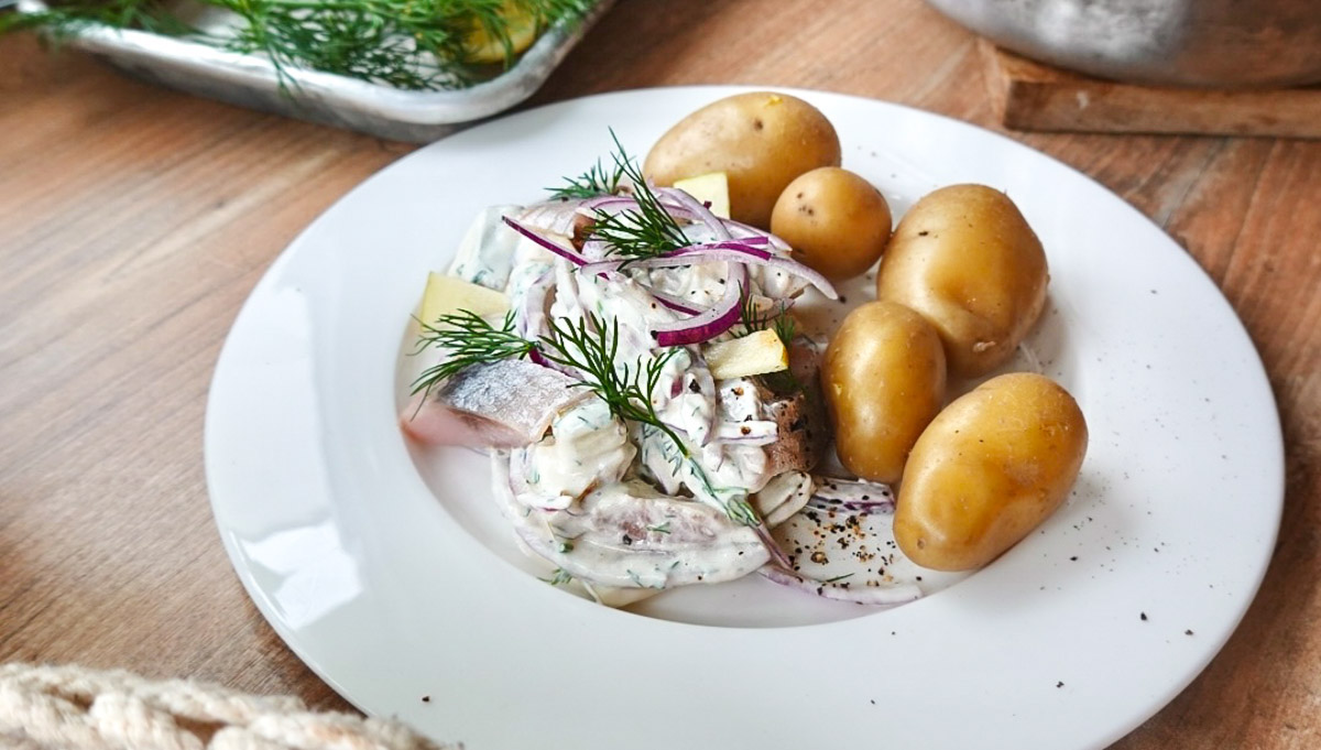 Teller mit Sahnehering, roten Zwiebeln, Dill und kleinen Kartoffeln auf einem Holztisch.