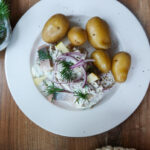 Teller mit Hering in Sahnesoße, roten Zwiebeln, Apfelstücken und Kartoffeln, garniert mit Dill auf Holztisch.