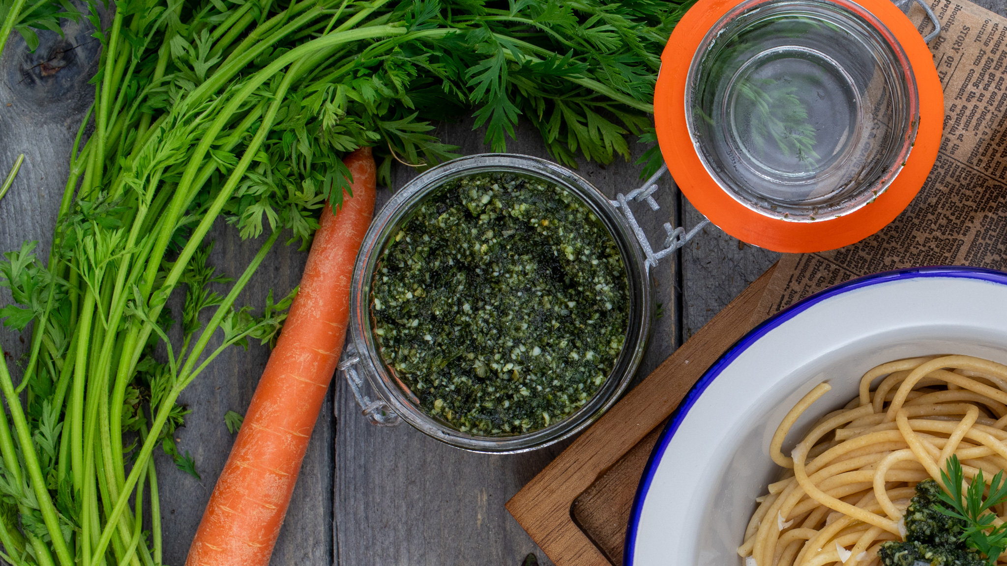 Ein Glas grünes Pesto neben einer Karotte und Karottengrün auf Holz. Rechts darunter ein Teller Spaghetti mit Pesto.