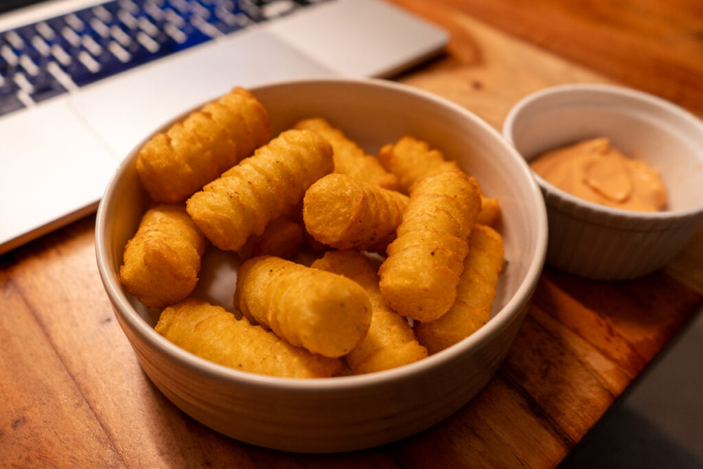 Schale mit Kartoffelkroketten und Schale mit Dip auf Holztisch, Laptop im Hintergrund.