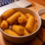 Schale mit geriffelten Kartoffelstücken und Schälchen mit Dip auf Holztisch neben Laptop.