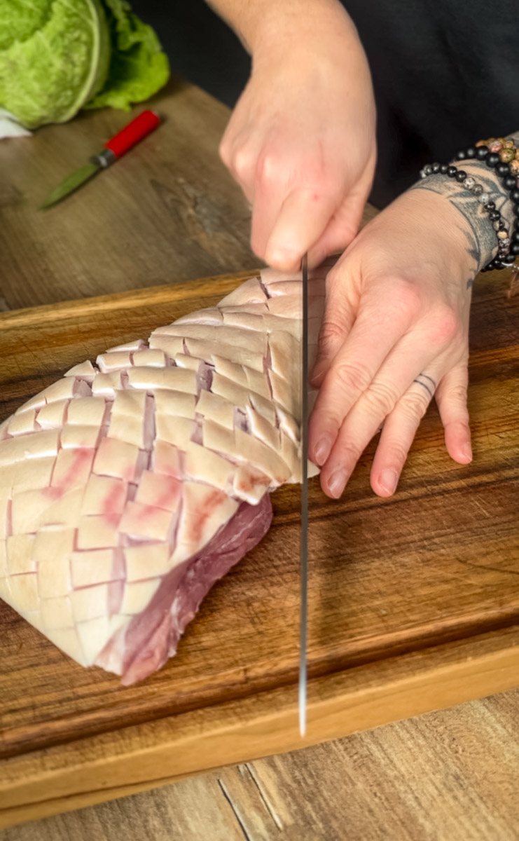 Krustenbraten-aus-Schweinebauch-aus-der-Heissluftfritteuse-02 Hand schneidet mit einem Messer ein gekreuzt eingeschnittenes Stück Schweinefleisch auf einem Holzbrett.