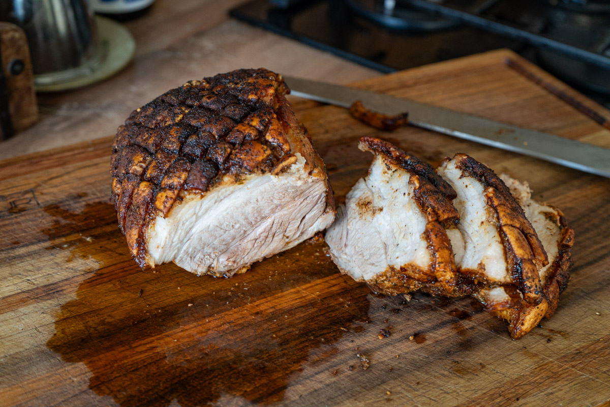 Krustenbraten-aus-Schweinebauch-aus-der-Heißluftfritteuse-01 Gebratenes Schweinefleisch mit knuspriger Kruste auf Holzbrett, teilweise aufgeschnitten, daneben ein Messer.
