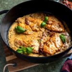 Hähnchen in cremiger Sauce mit Basilikum und getrockneten Tomaten in einer schwarzen Pfanne auf Holzbrett serviert.