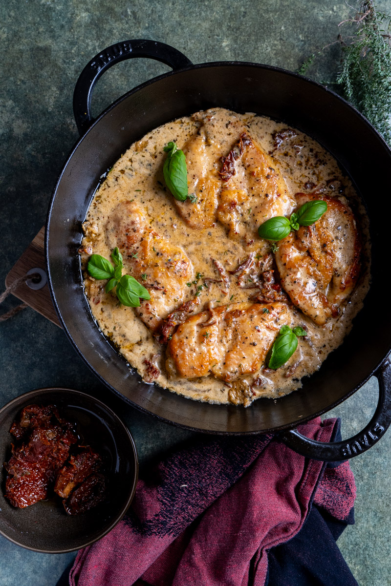 Marry-Me-Chicken-04 Hähnchenbrustfilets in cremiger Soße mit Kräutern und getrockneten Tomaten, garniert mit Basilikumblättern in schwarzer Pfanne.