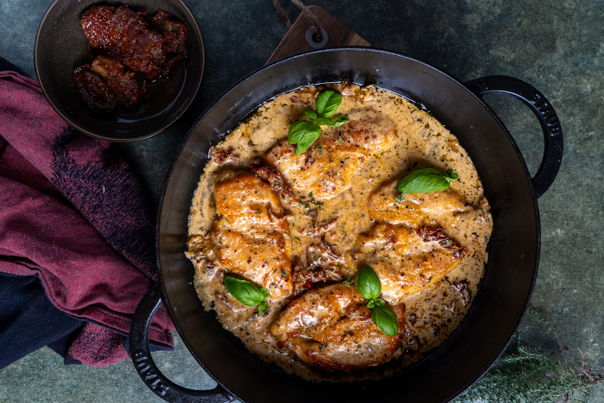 Marry-Me-Chicken-5 Hühnchen in cremiger Sauce mit Basilikum, serviert in einer schwarzen Pfanne, daneben getrocknete Tomaten in einer Schale.