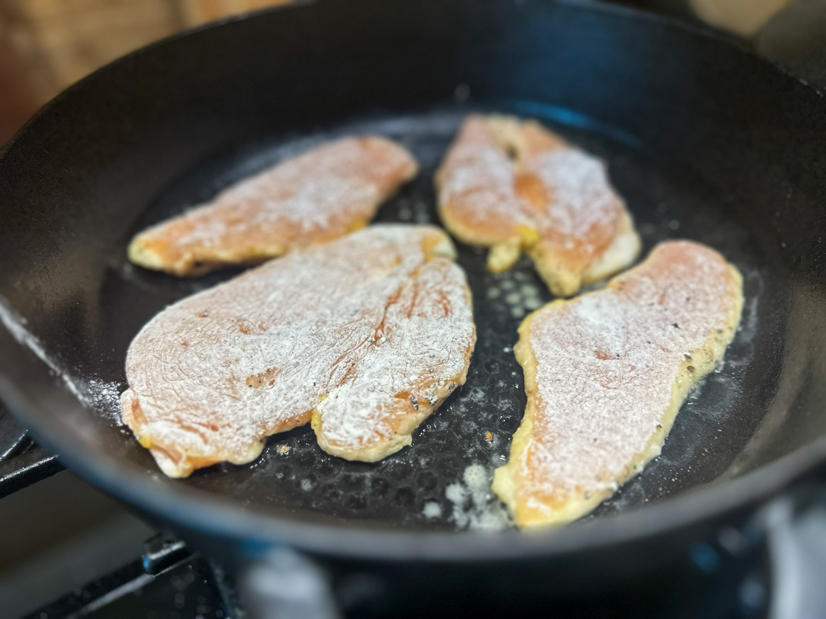 Marry-Me-Chicken-03 Vier mehliert panierte Hähnchenschnitzel braten in einer schwarzen Pfanne.