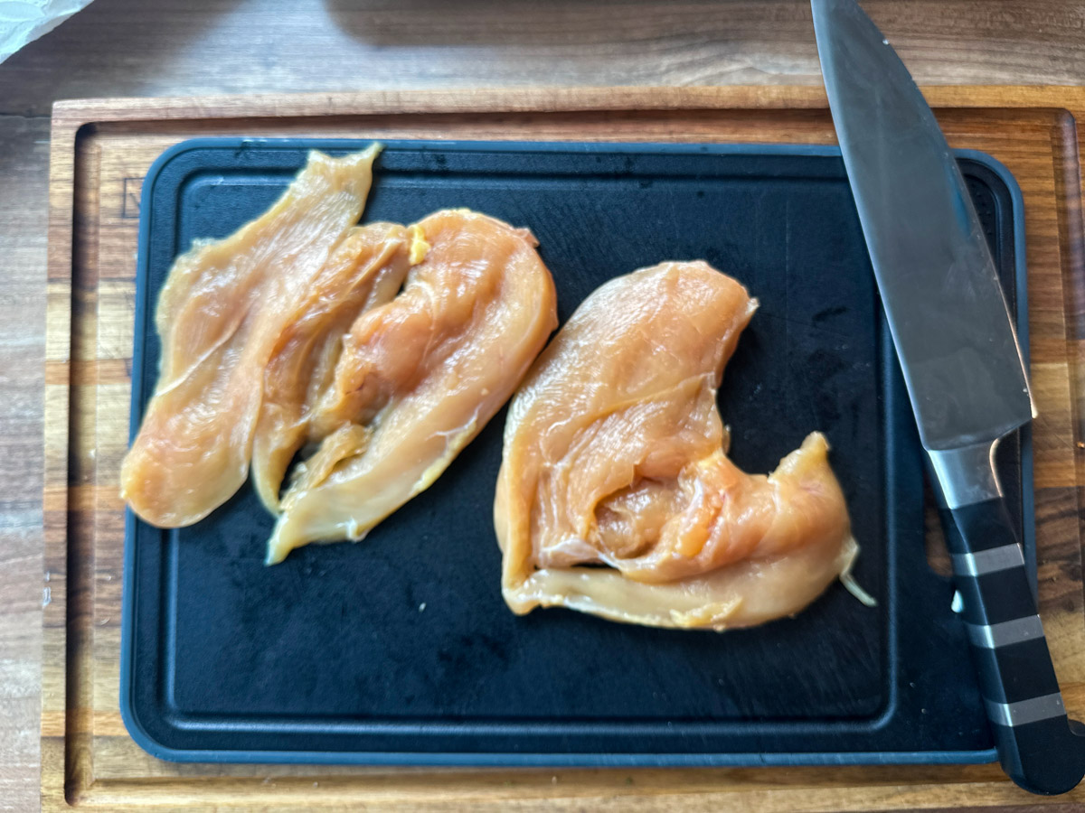 Marry-Me-Chicken-02 Zwei rohe Hähnchenbrustfilets auf einem schwarzen Schneidebrett mit Messer.