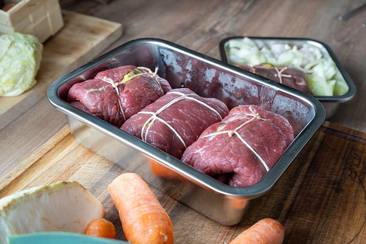 Rinderrouladen in einer Metallschale, umgeben von Karotten und Kohl auf einem Holztisch.